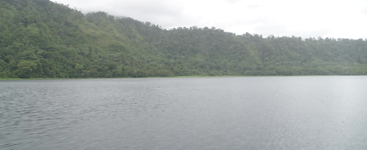 Raindrops fall on a tranquil lake surrounded by a lush green forested mountain on an overcast day