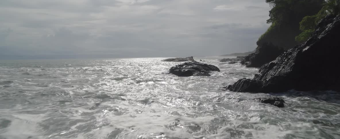 Slow aerial push-in over a shimmering, rocky ocean coastline on a bright but overcast day
