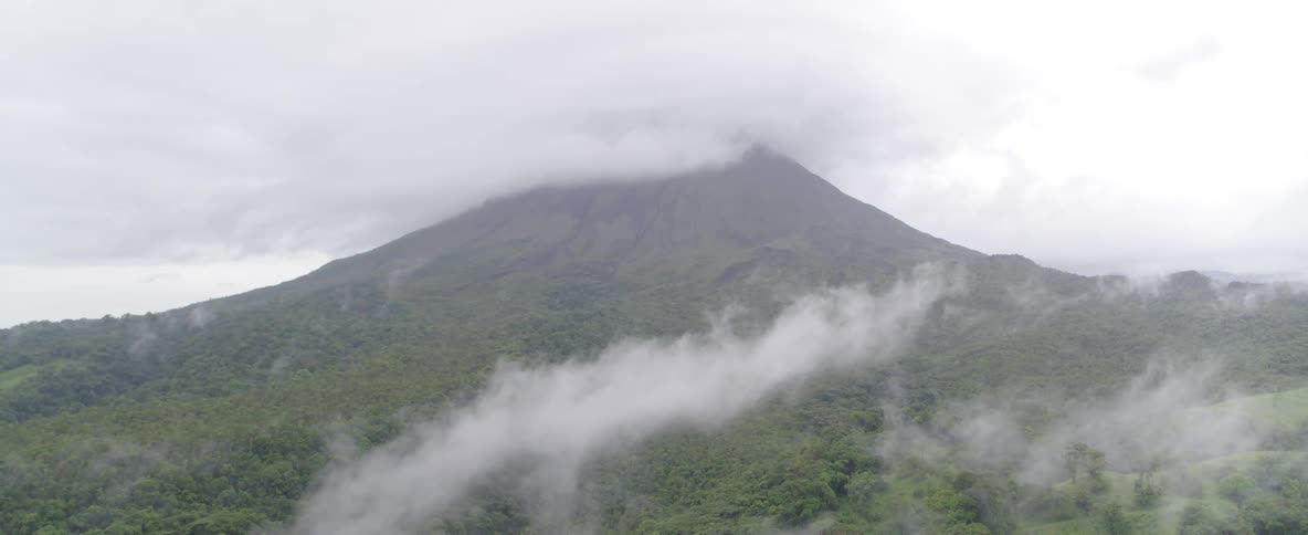 Clouds and fog drift across the forested slopes of a massive volcano on an overcast day
