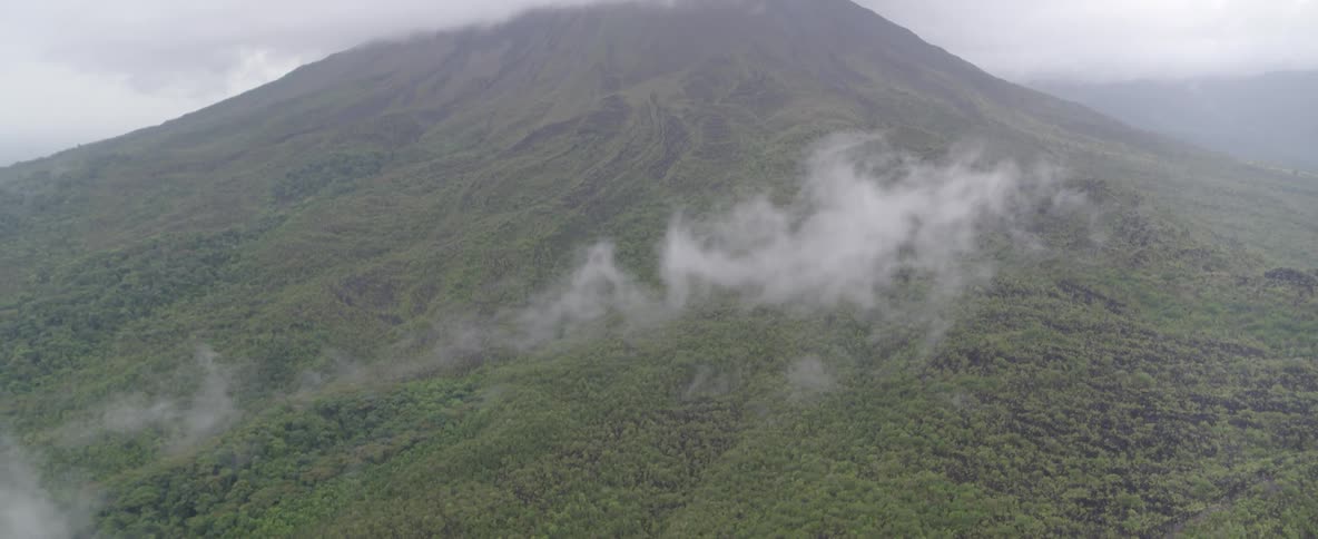 An aerial tilt-down from a majestic volcano shrouded in clouds to the vast, lush green forest at its base
