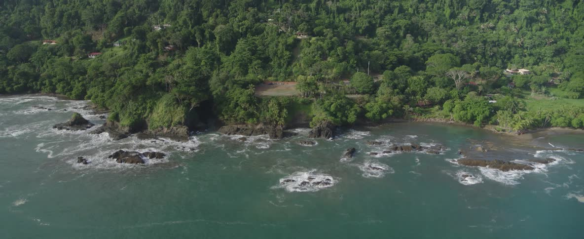 An aerial view of a rugged, tropical coastline where a dense jungle meets the rocky shore and ocean waves