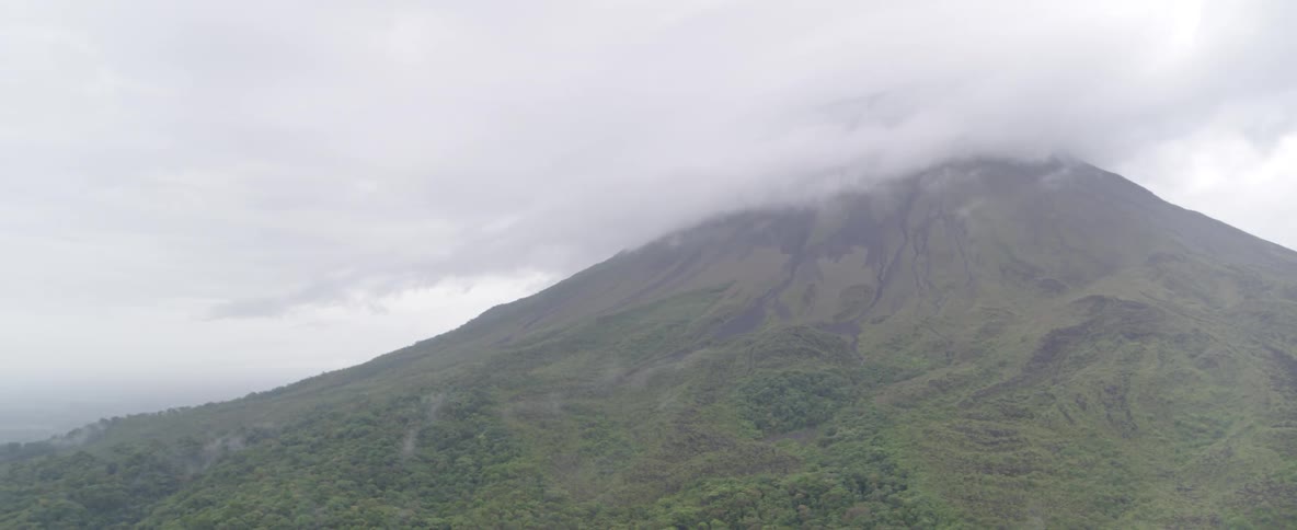 Slow aerial pan away from a cloud-covered volcano peak over a vast green rainforest on a misty day
