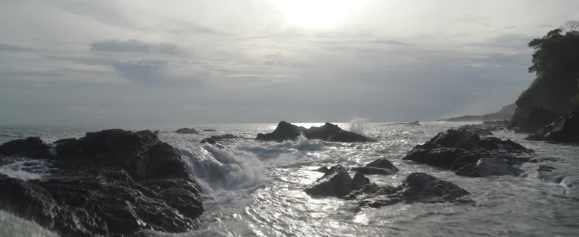 An aerial shot slowly glides over a rocky shoreline as waves gently crash and the sun glimmers on the water's surface