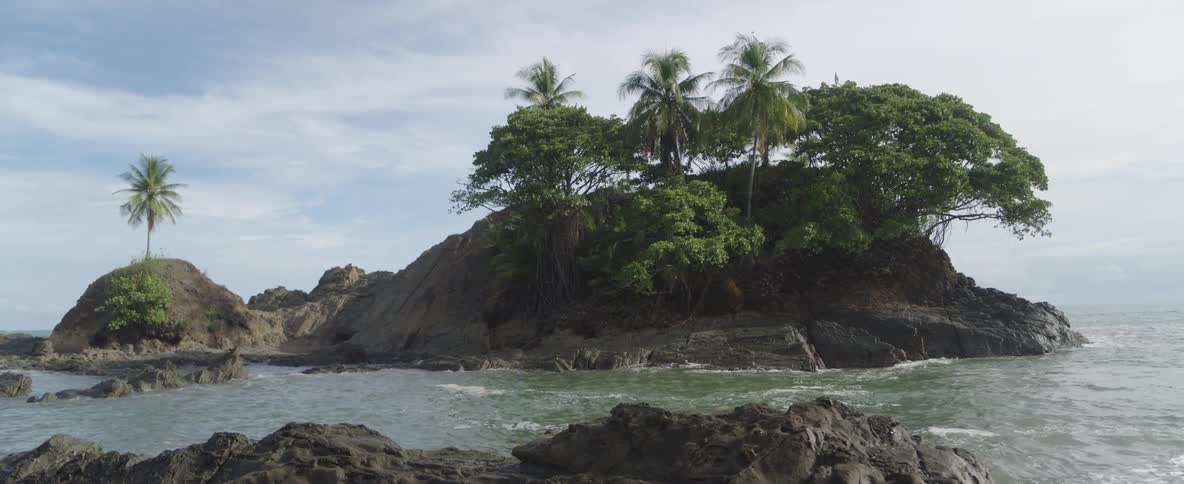 A small, rocky tropical island with lush palm trees sits just off the coast as gentle waves lap at its base