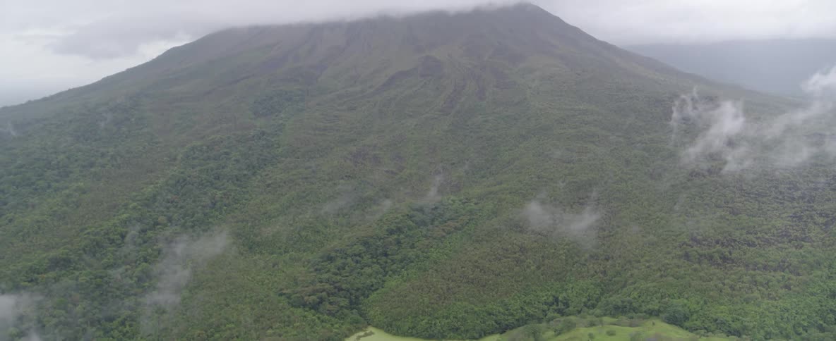 An aerial shot pans up from a small lake to reveal a massive, cloud-covered volcano surrounded by lush green forest
