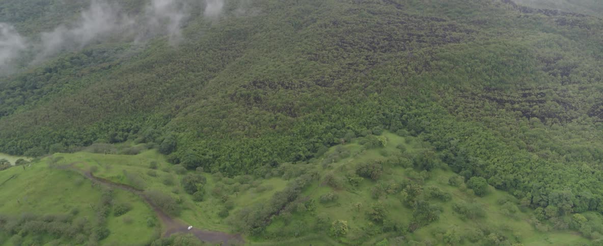 An aerial view tilts up from a small road in a valley to reveal a massive, lush green mountain covered in dense forest and shrouded in clouds