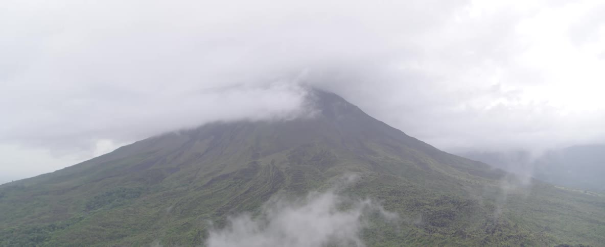A serene and moody wide shot captures a large volcano covered in lush vegetation, with clouds obscuring its summit