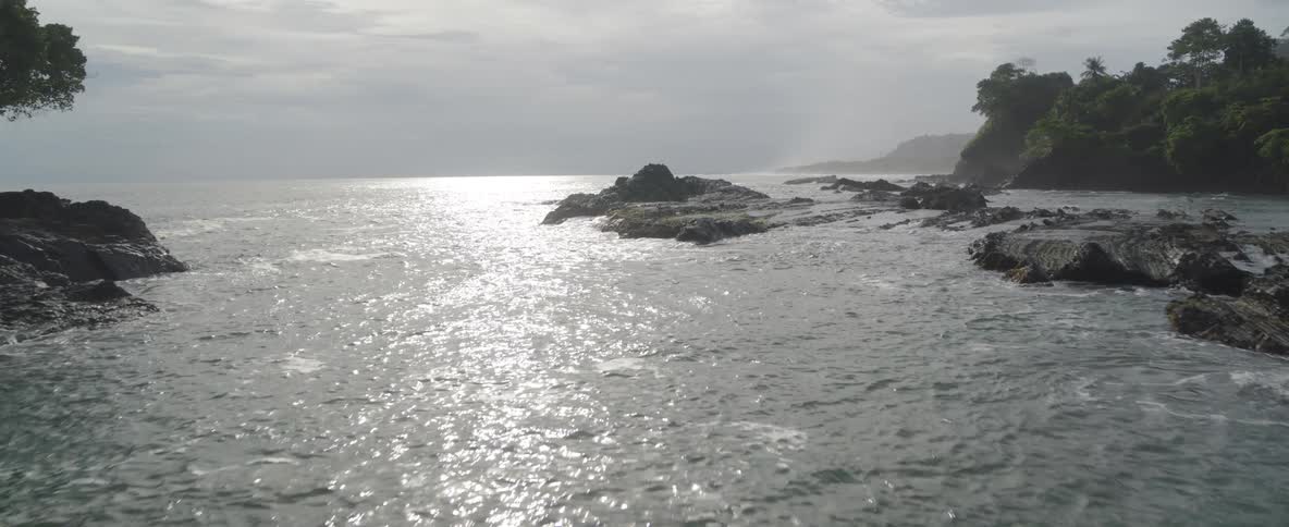 The sun glitters on the surface of the ocean as it gently washes over a rocky coastline on an overcast day