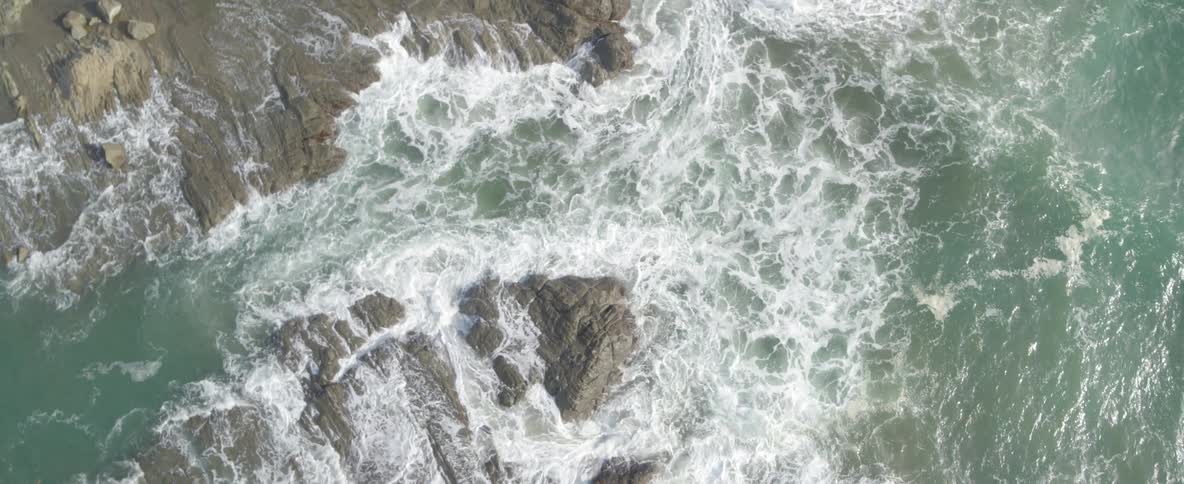 Slow-motion aerial top-down view of powerful ocean waves crashing over a rocky shoreline