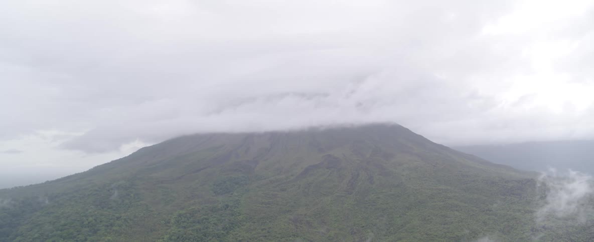 A static, wide shot shows a lush green volcano with its peak shrouded in a thick layer of clouds on a grey, overcast day