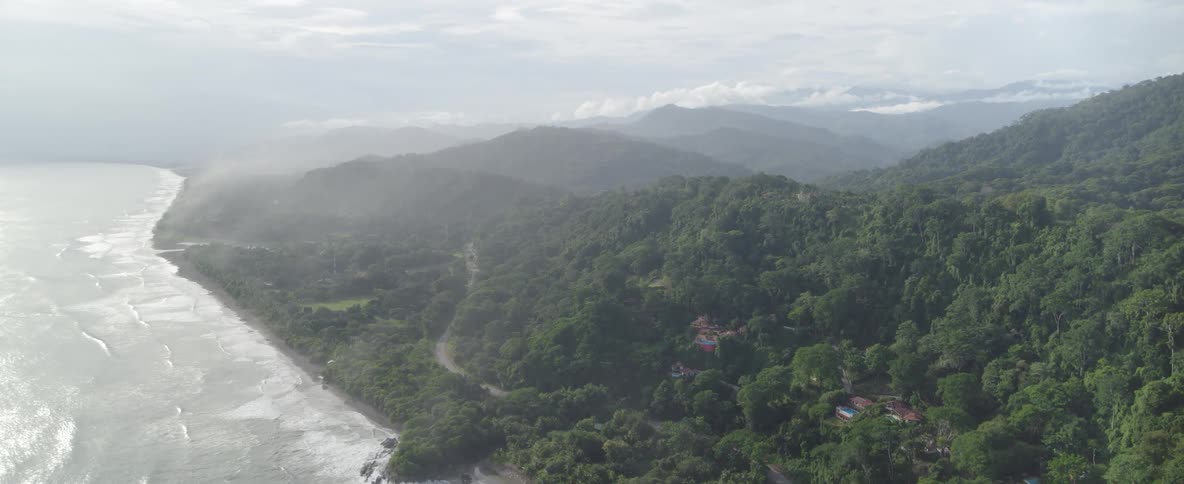 A slow aerial view pushes in over a misty tropical coastline where lush green mountains meet the ocean's gentle waves