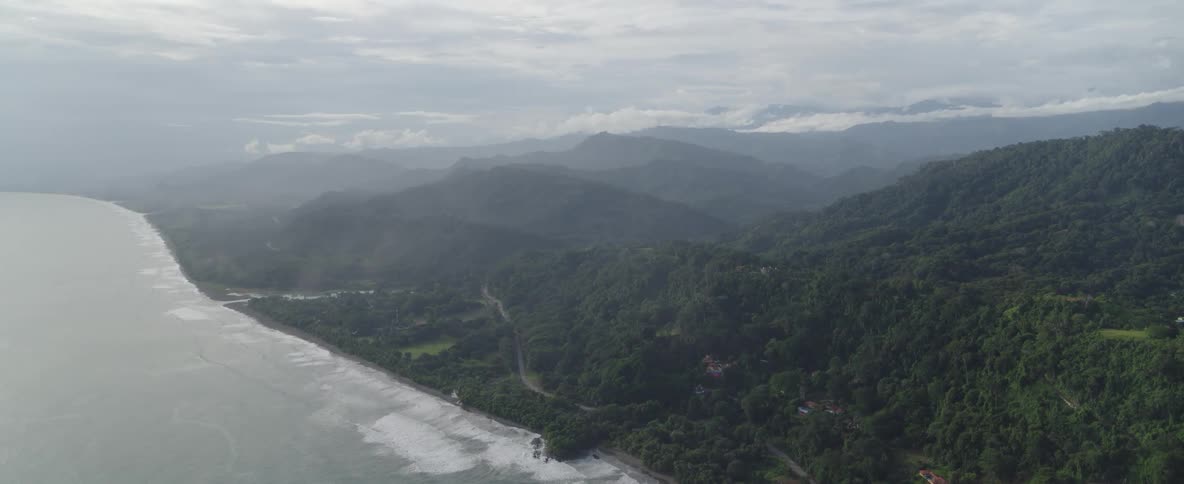 A wide aerial shot captures the lush tropical coastline where the jungle-covered mountains meet the ocean on a hazy day