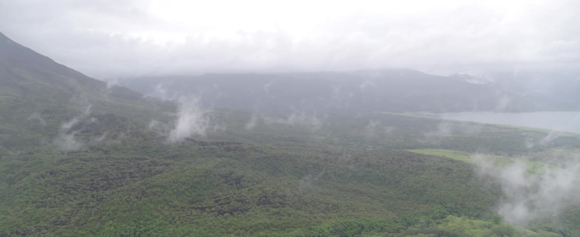 Slow aerial pan over a foggy mountain range reveals a lush green valley, a distant lake, and a large volcano