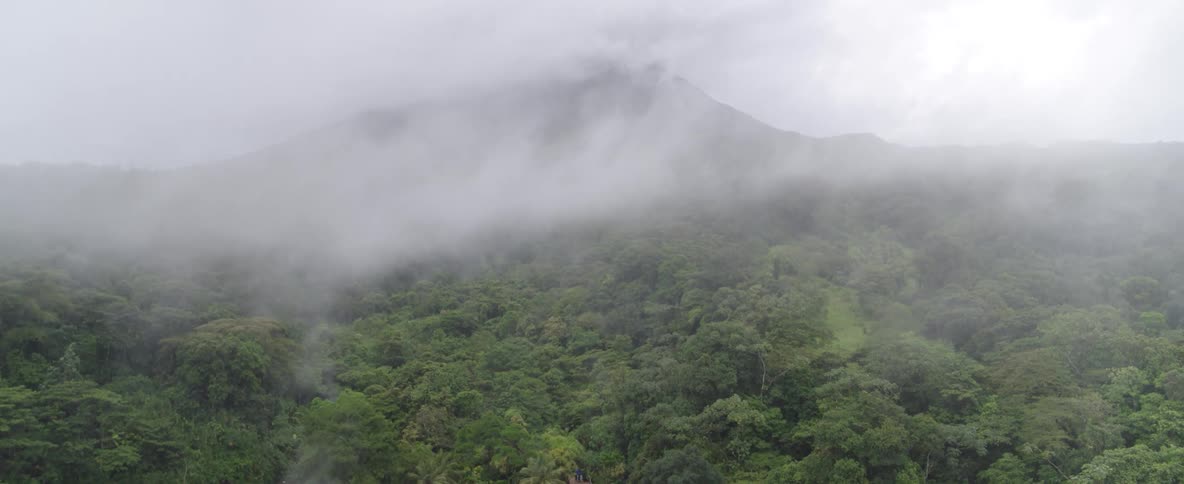 Aerial view flying backwards over a dense, foggy rainforest to reveal a large volcano shrouded in clouds