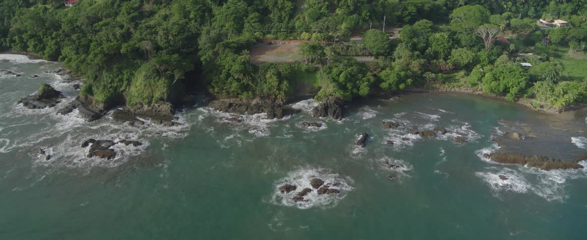 A slow aerial pullback reveals a vast, dense tropical forest behind a beautiful rocky coastline