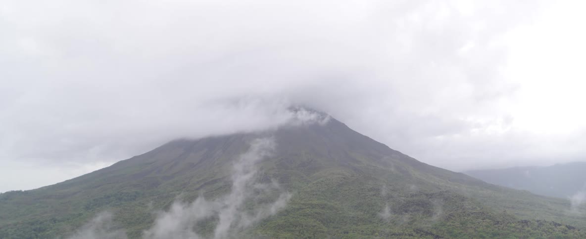 A wide, static aerial view shows a majestic volcano covered in a lush green forest, with its peak shrouded in thick clouds and mist