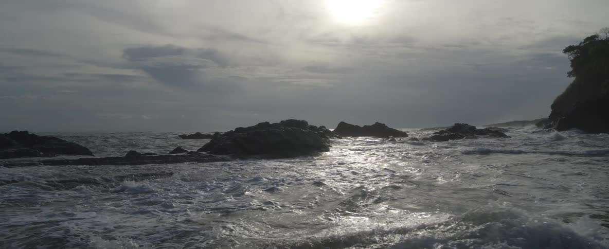 Dramatic slow-motion view of ocean waves crashing over rocks with the sun reflecting on the water's surface