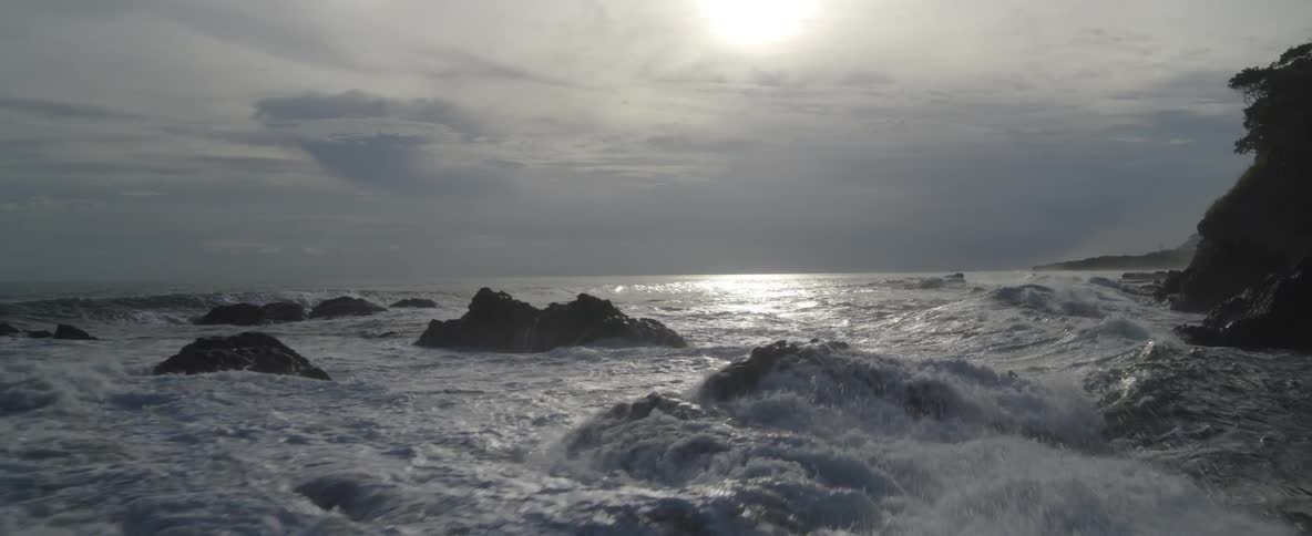 Slow motion wide shot of the ocean with waves crashing over rocks as the sun reflects on the water