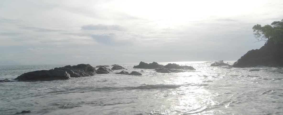 A slow pull-back shot reveals a tranquil, rocky coastline as gentle waves wash ashore under a bright, overcast sky
