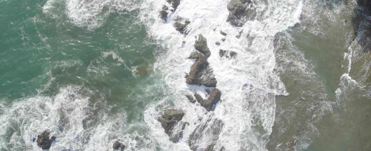 A top-down aerial shot captures the power of ocean waves crashing onto a rugged, rocky shore, transitioning with a dynamic spin before ascending