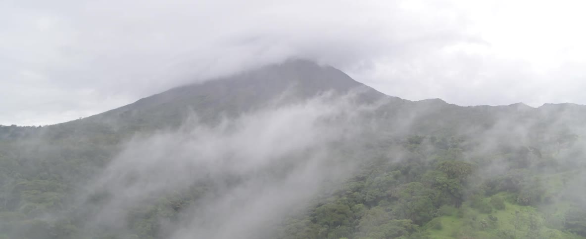 An aerial drone shot flies slowly through thick fog to reveal a majestic volcano surrounded by a lush green jungle