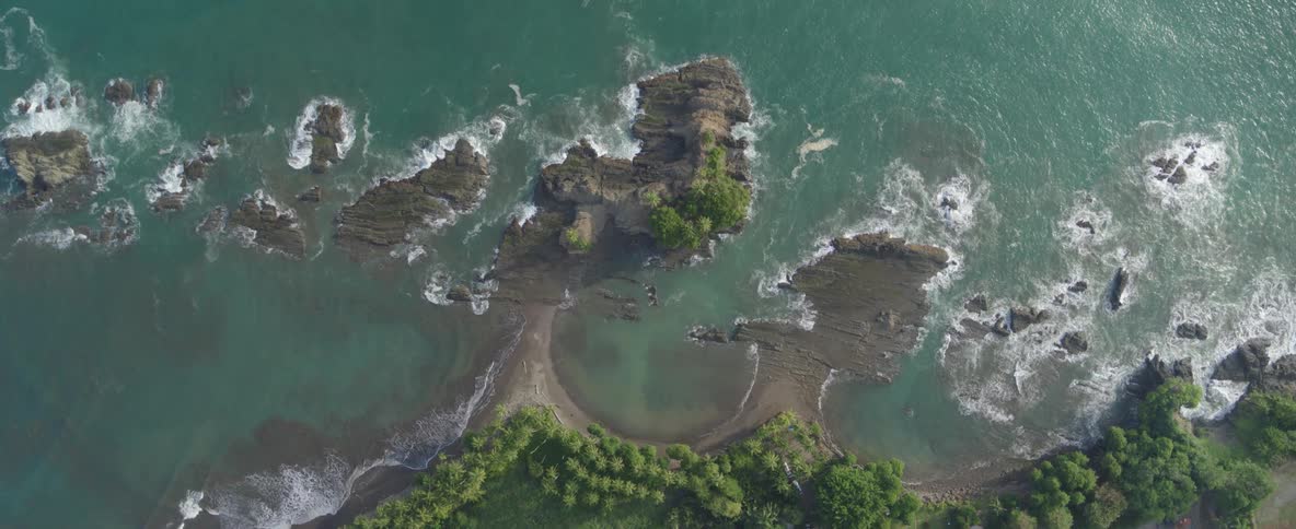 An aerial top-down view slowly pulls back to reveal a stunning tropical coastline with rocky shores, a hidden sandy cove, and lush green jungle