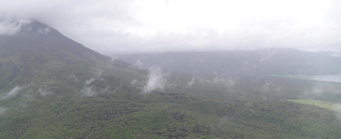 A misty aerial view pans across a vast green valley and a distant lake to reveal a cloud-capped mountain