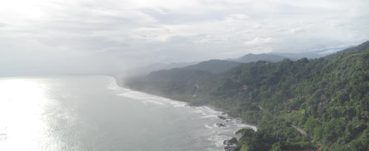 A serene aerial view of a misty, jungle-covered coastline where lush mountains meet the sea on a cloudy day