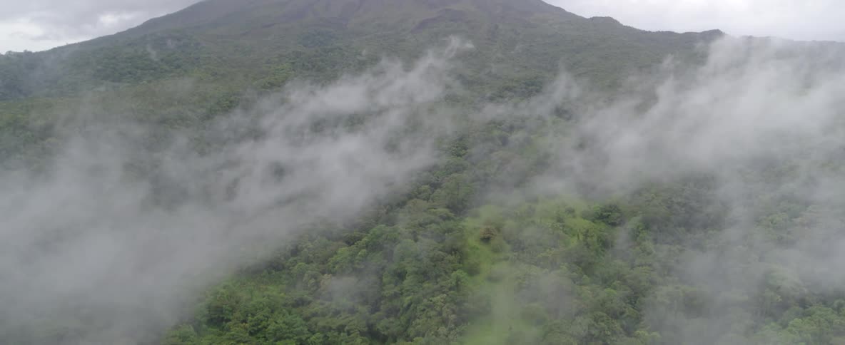 Aerial view tilts down from a large volcano peak covered in clouds to the dense, misty rainforest below