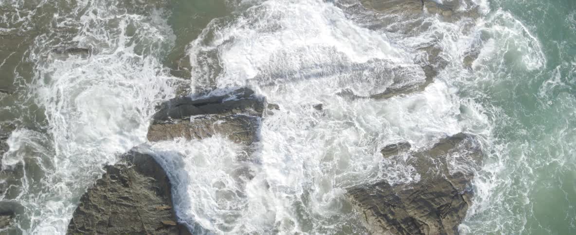 A slow-motion, top-down aerial view of powerful ocean waves crashing over rugged coastal rocks