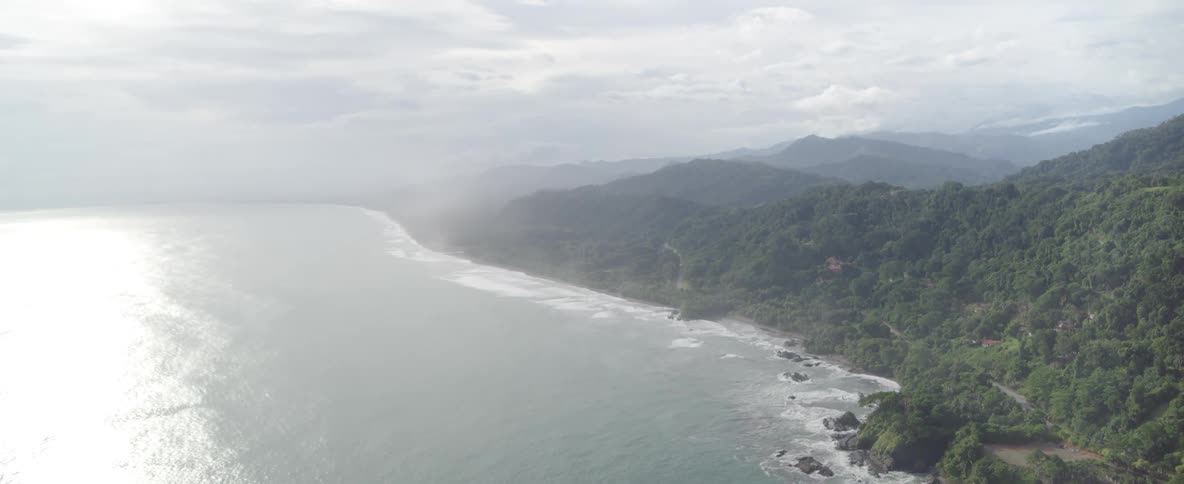 A stunning aerial view of a misty tropical coastline with lush green mountains and the sun reflecting on the ocean