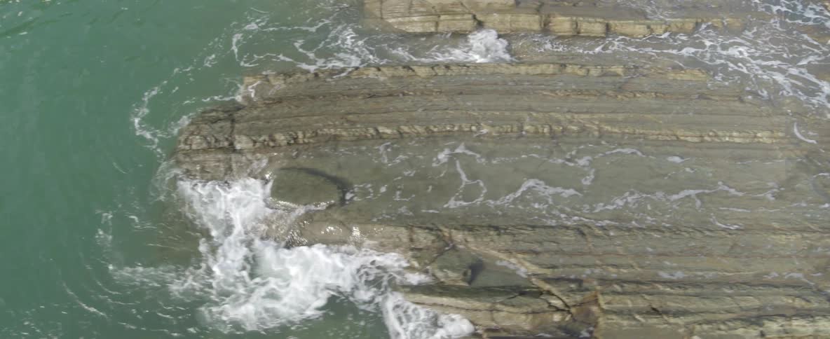 An aerial top-down view shows ocean waves gently crashing against unique layered rock formations on a rugged coastline