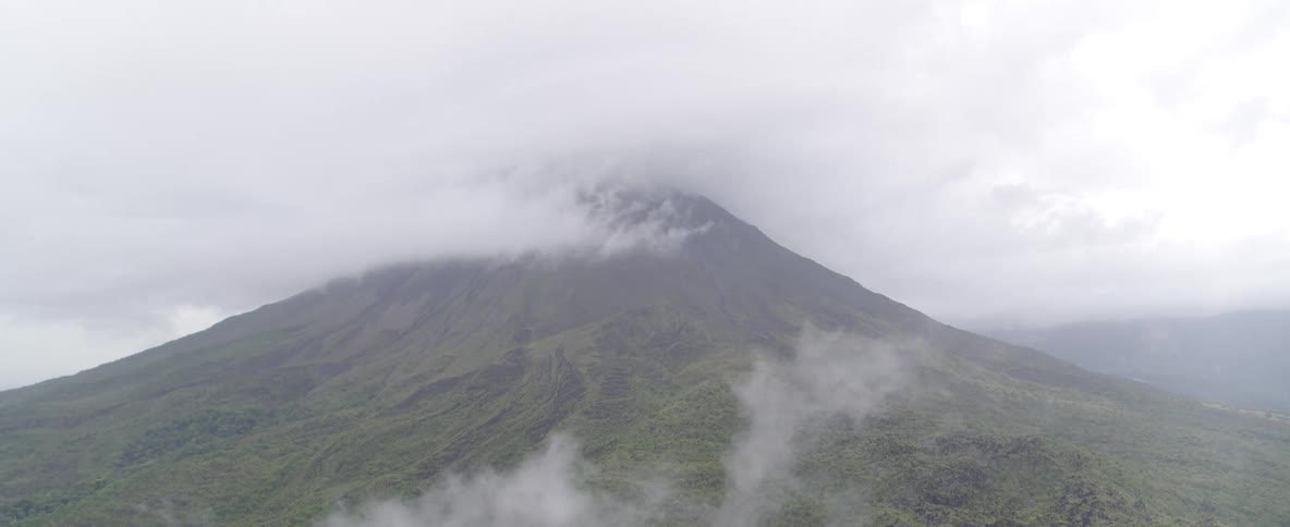 A majestic, green volcano stands tall as its peak is enveloped in a thick blanket of clouds and fog
