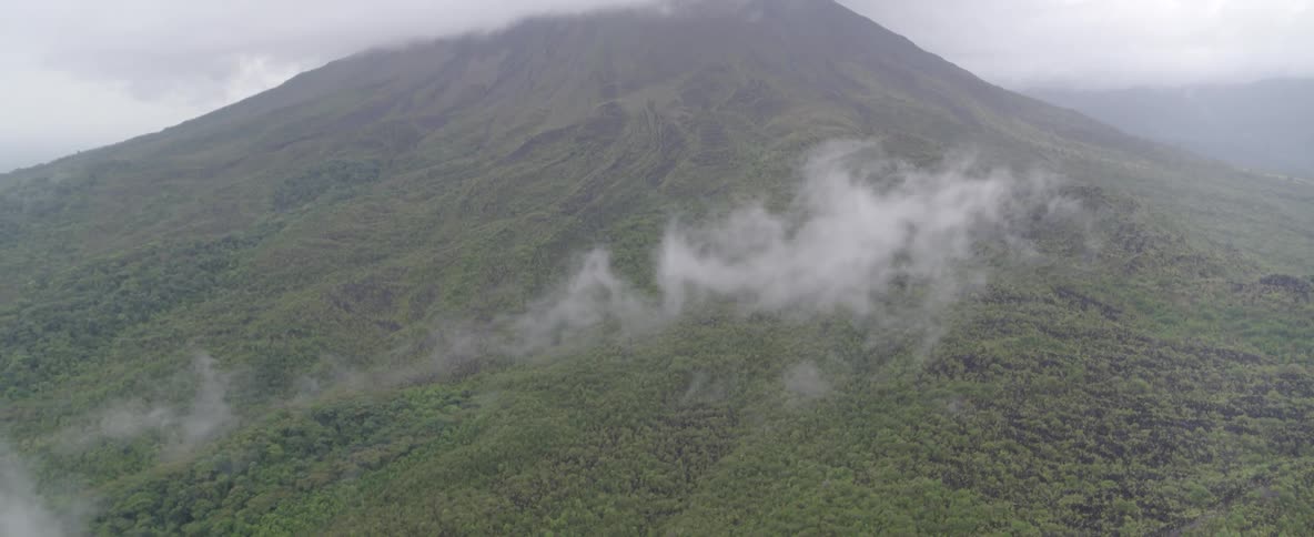 An aerial view of a majestic green volcano shrouded in mist, tilting down to reveal a lush rainforest and valley below