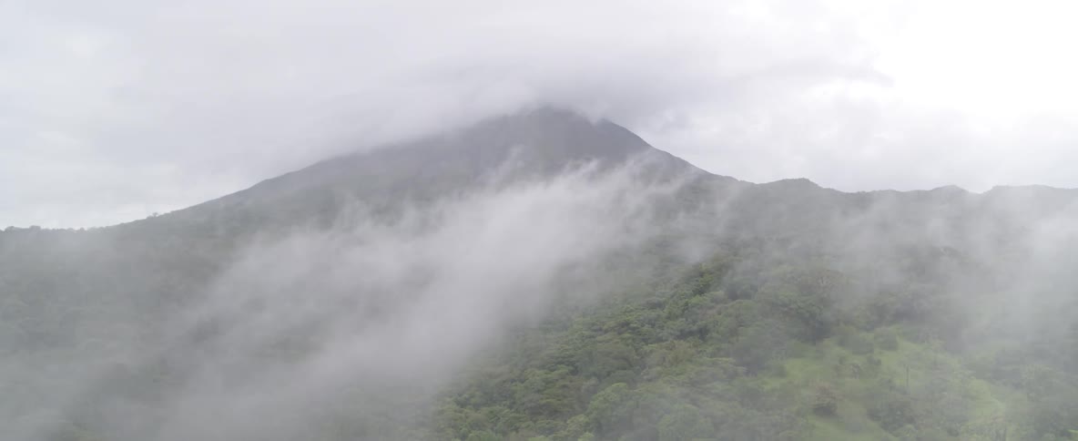A drone flies slowly through the misty clouds revealing a majestic, forest-covered volcano on an overcast day
