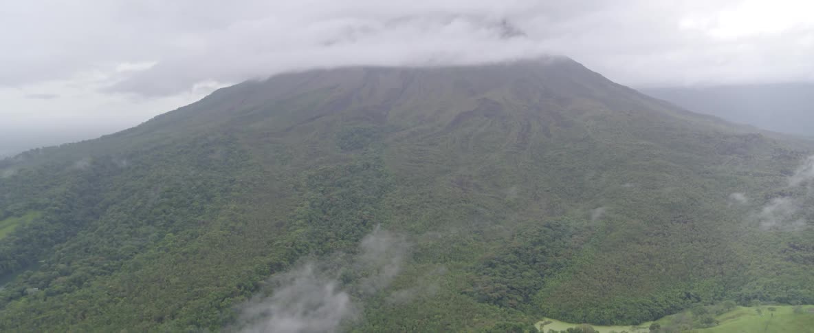 A dramatic aerial shot tilts down from a majestic, cloud-covered volcano to the sprawling tropical rainforest at its base