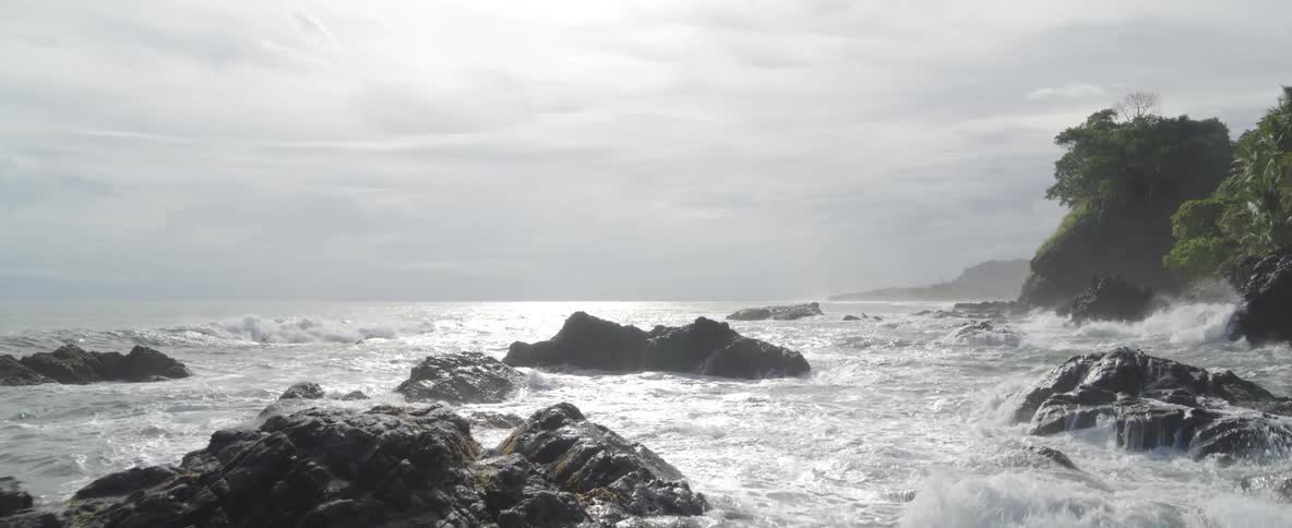 Ocean waves crash over dark, volcanic rocks in dramatic slow motion along a tropical coastline
