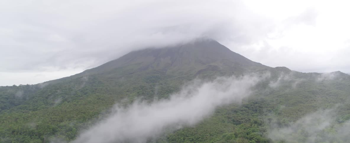 A static aerial view of a majestic volcano shrouded in thick clouds and mist over a lush green forest