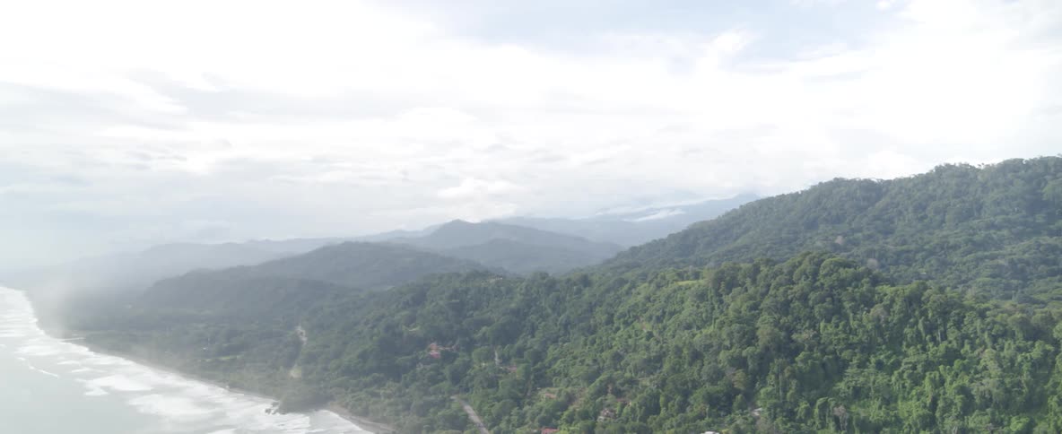 An expansive aerial view pans out from dense jungle-covered mountains to reveal the vast ocean coastline