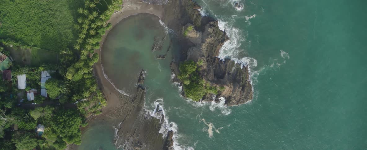 An aerial top-down shot slowly rises to reveal a stunning tropical coastline with a rocky island and secluded cove