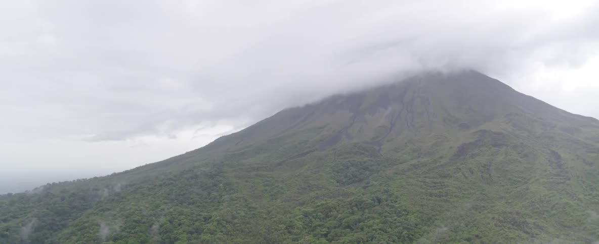 A slow aerial pan reveals a massive green volcano with its peak hidden by thick clouds and fog
