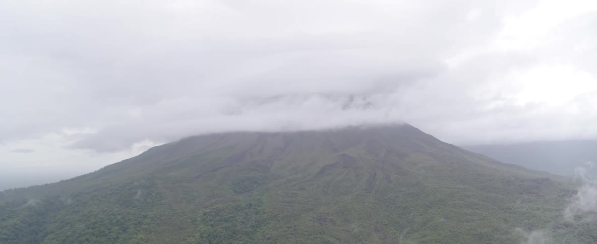 A slow tilt-down shot reveals the expansive green jungle at the base of a majestic, cloud-shrouded volcano