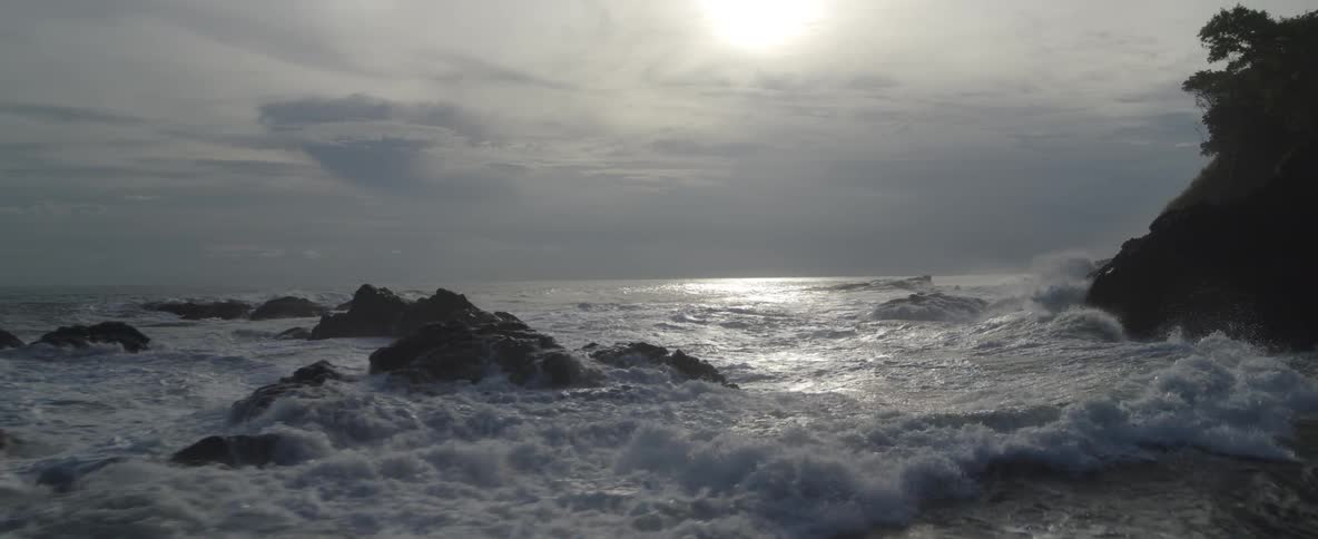 A powerful slow-motion shot of ocean waves crashing onto a rocky coastline as the sun glints off the water's surface