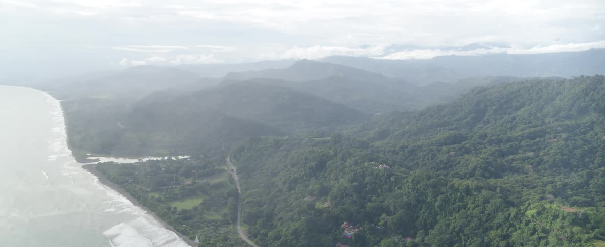 An expansive aerial pullback reveals a winding coastal road separating a sandy beach from the lush, misty jungle mountains
