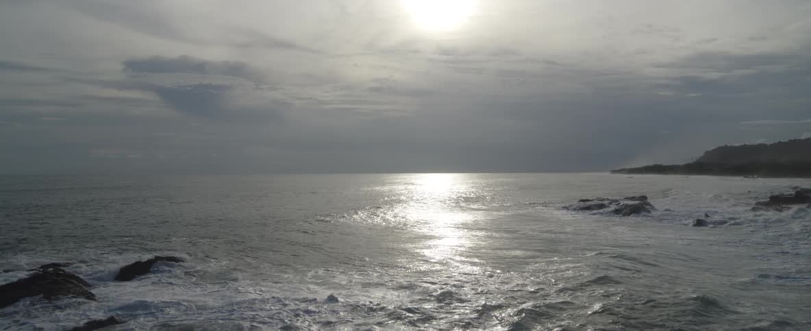 An aerial shot slowly rises over a rocky ocean coastline, revealing the vast sea with the sun's bright reflection on the water