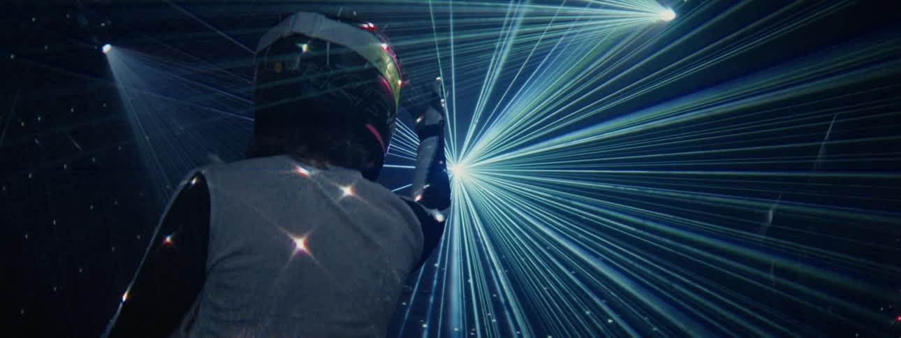 A person wearing a futuristic helmet stands in a dark room filled with a dazzling laser light show