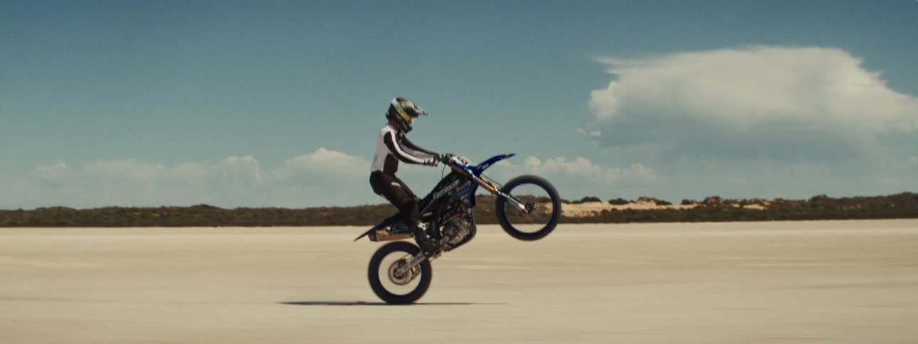 A profile shot captures a skilled motocross rider performing a sustained wheelie on a dirt bike across a vast desert landscape