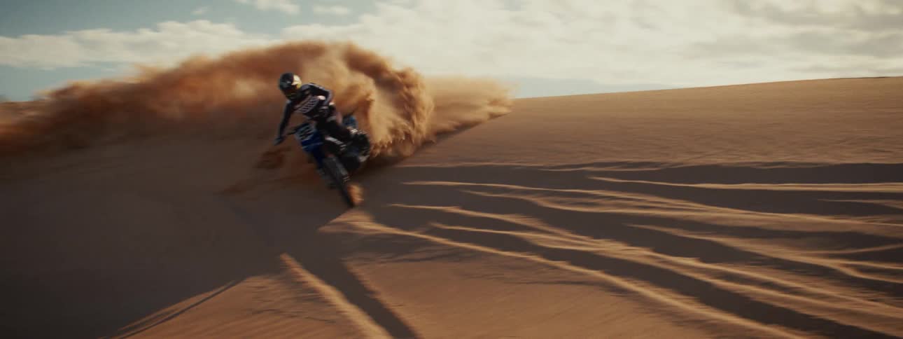 A motocross rider carves a turn on a coastal sand dune, kicking up a huge cloud of sand in slow motion
