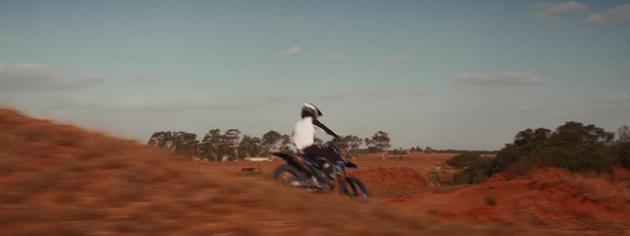A motocross rider speeds along a red dirt track in slow motion, kicking up a massive cloud of dust while making a sharp turn