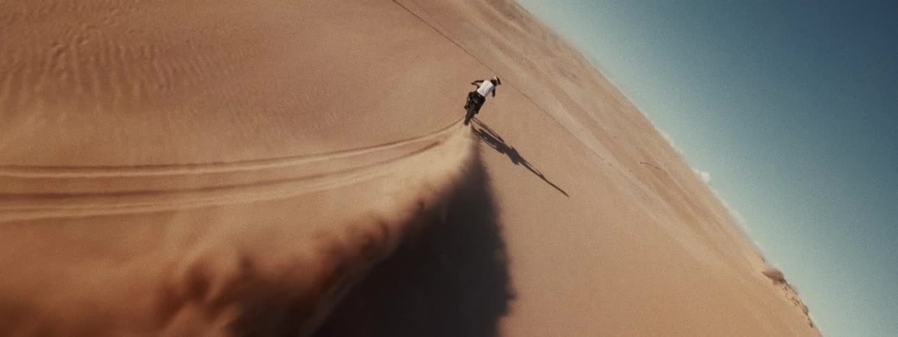 Cinematic slow-motion aerial view of a person riding a motorcycle down a steep desert sand dune